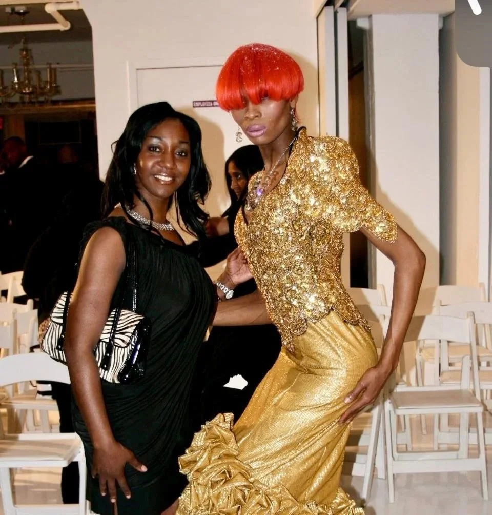 Two women posing at an indoor event, one in a black dress and the other in a gold, glittery gown with red hair.