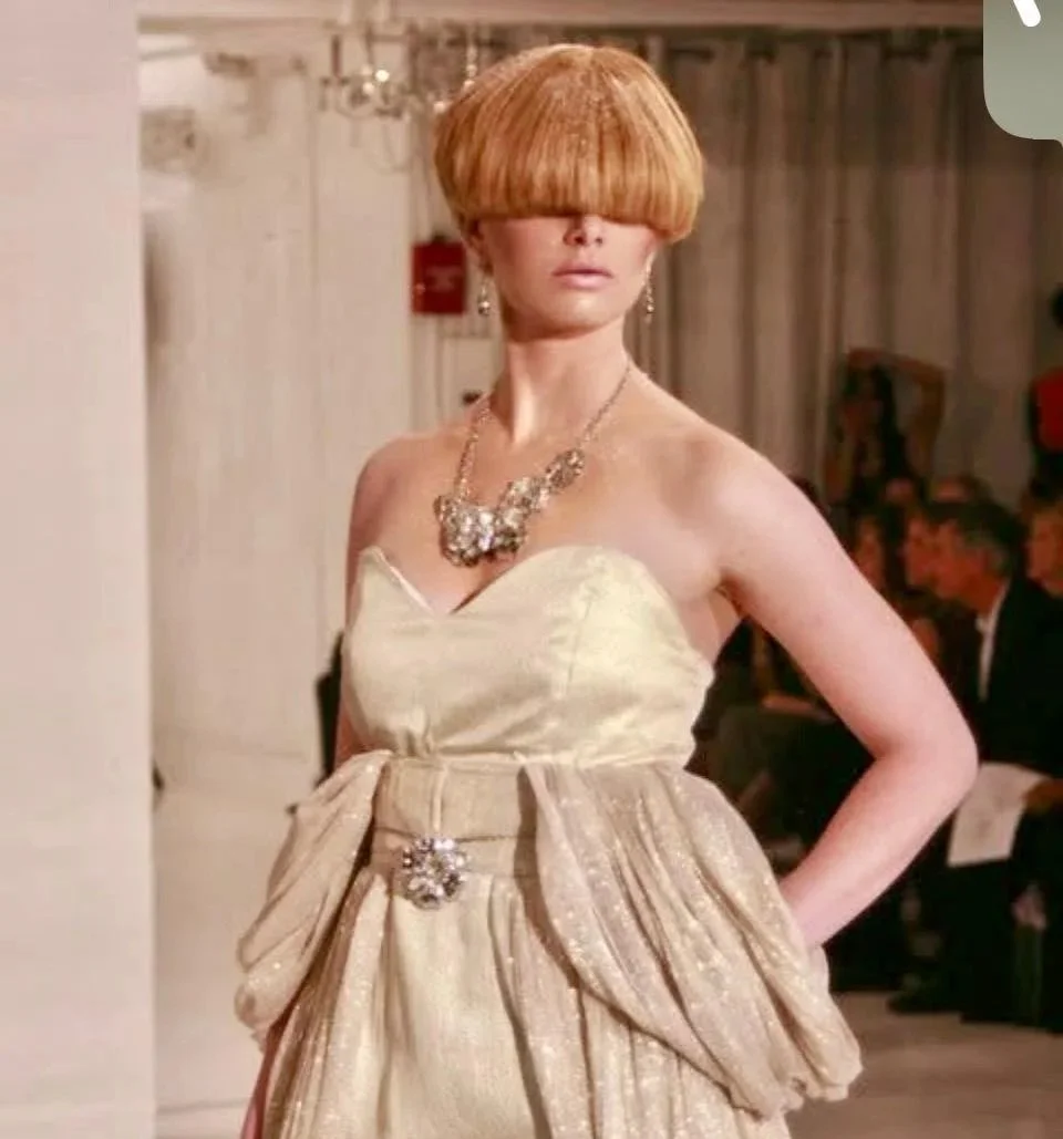 A woman in an elegant strapless beige gown with a ruffled skirt and large bow detail, accessorized with statement jewelry, standing in an indoor setting.