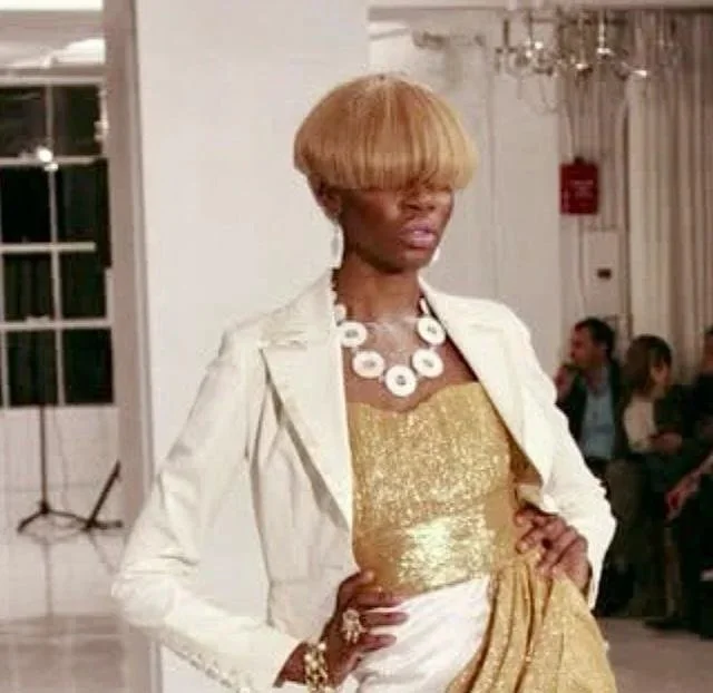 A woman with a large blonde bob haircut, wearing a gold dress, white blazer, and a chunky white necklace, posing with her hand on her hip at an indoor event.