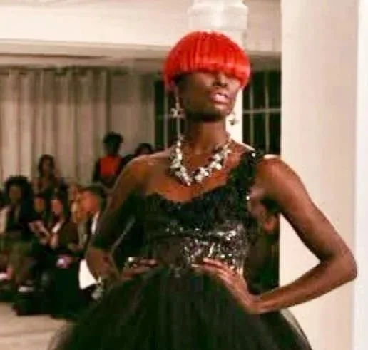 A woman with a short, bright red bob haircut wearing a black, sequined dress. She has jewelry, including earrings and a necklace, and stands confidently with her hand on her hip.