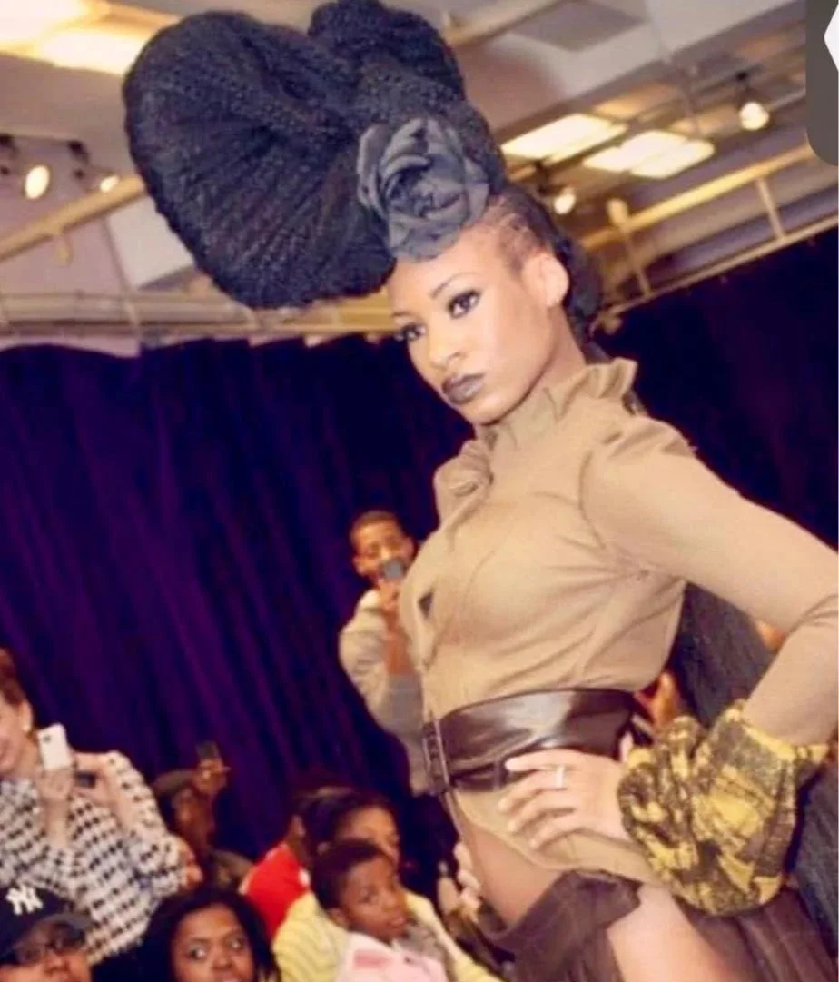 A woman with elaborate braided hair styled in an updo with a large bun and flower accessory, wearing makeup with dark lipstick, posing confidently with her left hand on her hip in front of an audience at an event.