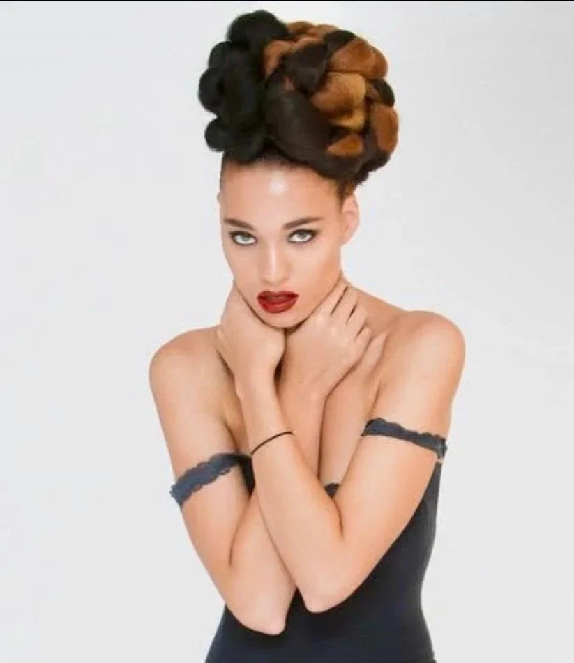 A woman with an elaborate hairstyle made of large braided buns in black and brown colors, wearing a black off-shoulder dress with lace trim, posing with her hands on her neck against a plain white background.