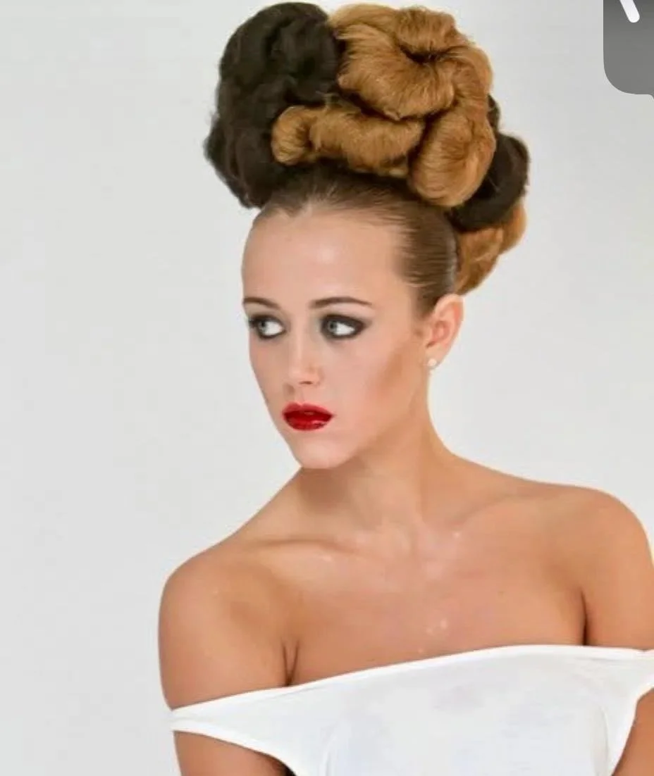 A woman with elaborate brown and black hair styled in large coils, wearing makeup with bold red lipstick, and a white off-shoulder top against a plain light background.
