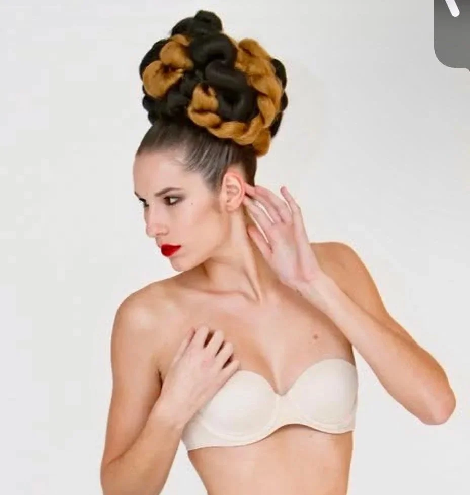A woman with an elaborate braided updo hairstyle, wearing a strapless beige bra, touching her ear with her right hand, against a plain white background.