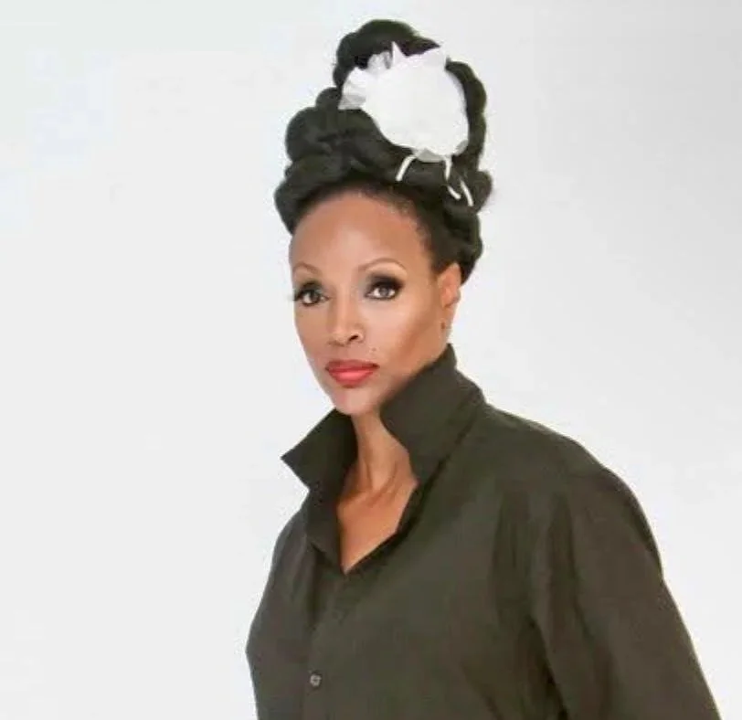 Woman with an elaborate hairstyle featuring a black bun with a white cloth wrapped around it, wearing a black button-up shirt with the collar turned up, against a plain white background.