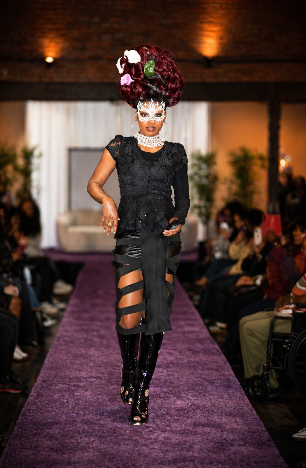 A model walking down a runway during a fashion show, wearing an elaborate outfit with a large, floral hairstyle, a decorative mask, a black lace top, and strappy black and leather skirt, with knee-high black patent boots.