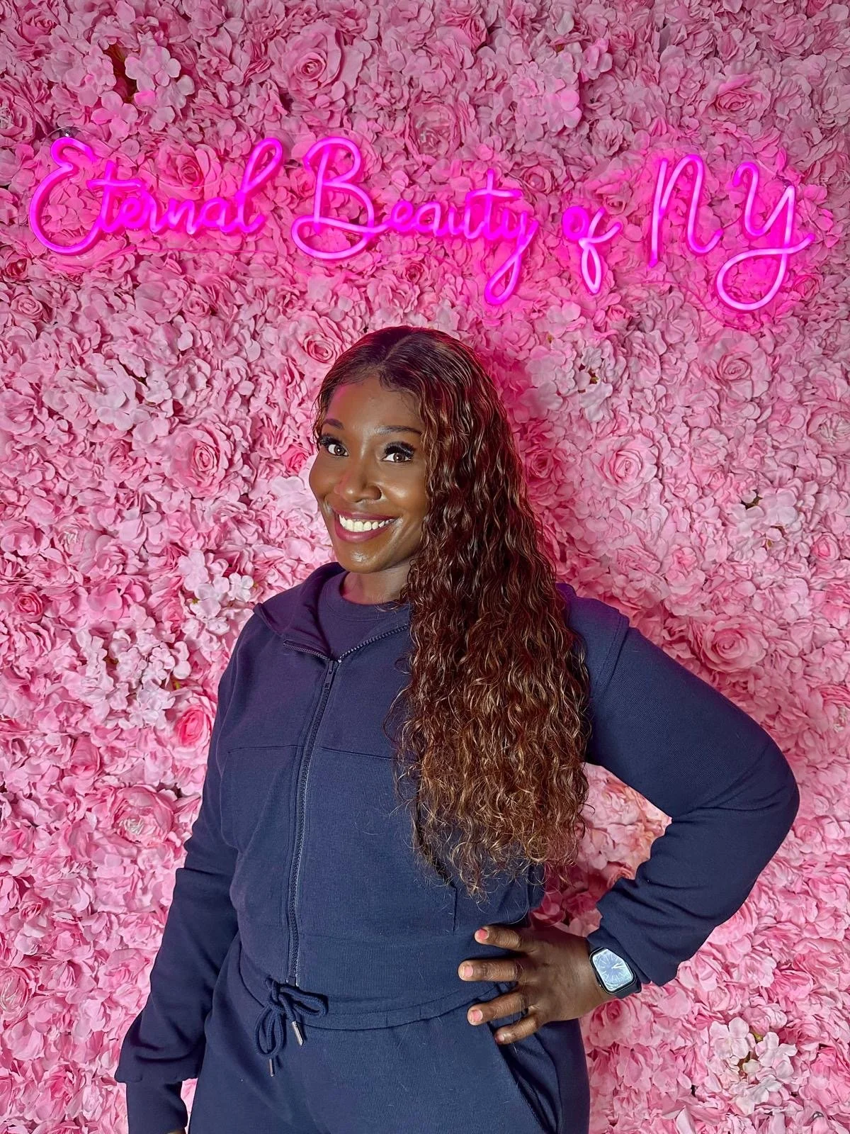A woman with long, wavy hair smiling, standing in front of a pink floral wall with a neon sign that reads 'Eternal Beauty of N'.