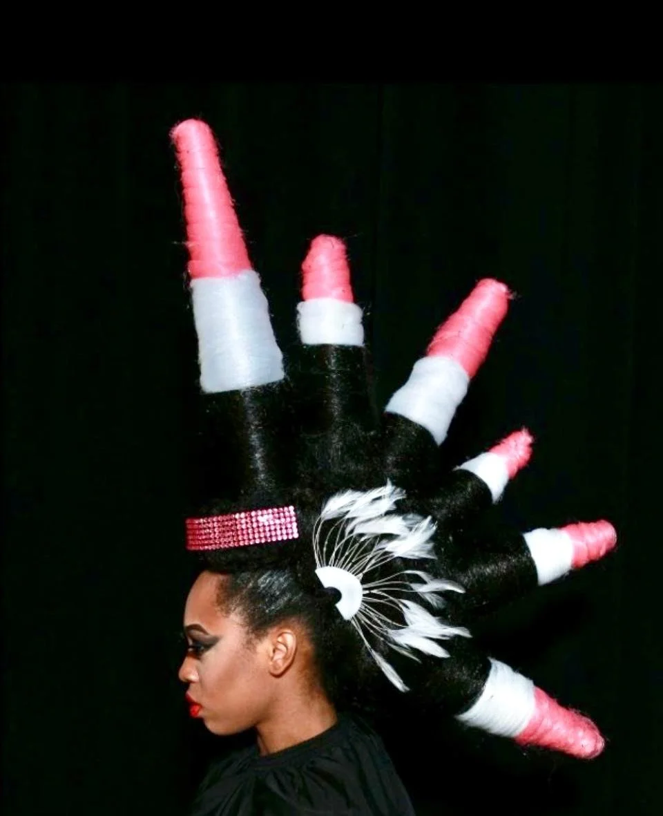 Side profile of a woman with an elaborate black, white, and pink feathered headdress resembling a hand with extended fingers decorated with pink, white, and black feathers.
