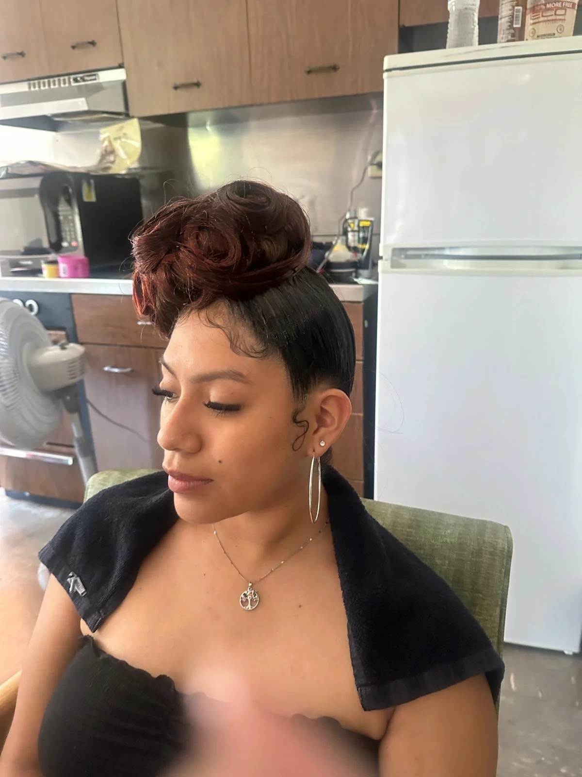 A woman with styled hair featuring a unique updo with waves and curls, wearing a black top and silver jewelry, sitting in a room with wooden cabinets, a white refrigerator, a fan, and kitchen appliances in the background.