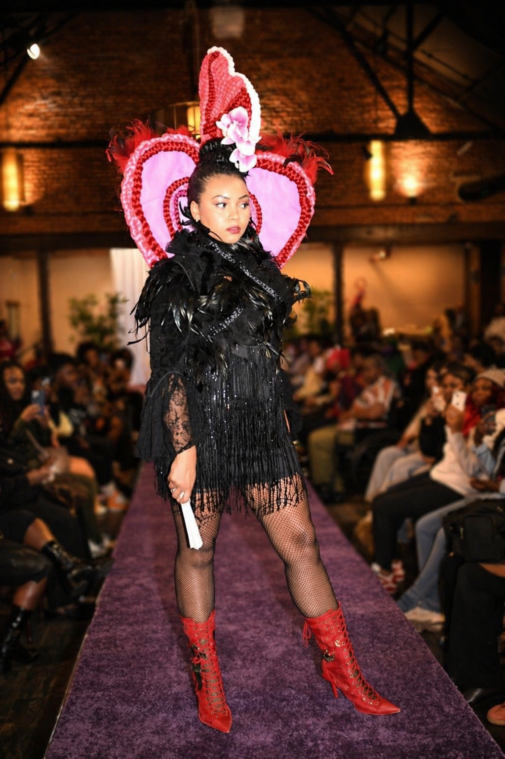 A woman fashion model walking down a runway at a fashion show, wearing a black fringed dress with lace details, black feathered accents, bright red lace-up high heel boots, and a large pink and red heart-shaped headpiece with flower decorations.