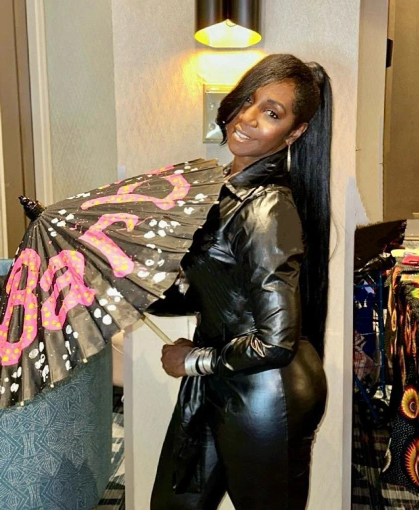 A woman with long black hair, smiling and wearing a shiny black leather outfit, holding a black umbrella with pink and yellow writing, standing indoors with warm lighting.