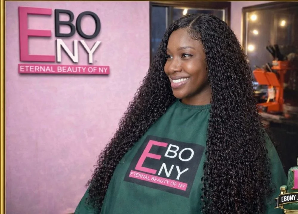 A woman with long, curly black hair smiling at a beauty salon or studio, wearing a green cape with the logo 'EBONY' and the tagline 'ETERNAL BEAUTY OF NY'. Behind her, there's a pink wall with the same logo and tagline.