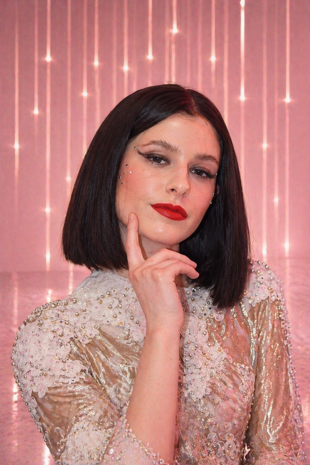 A woman with black shoulder-length hair and red lipstick poses with her hand near her chin against a pink backdrop with hanging light strands. She is wearing a sequined top decorated with floral appliqués and rhinestones around her eyes.