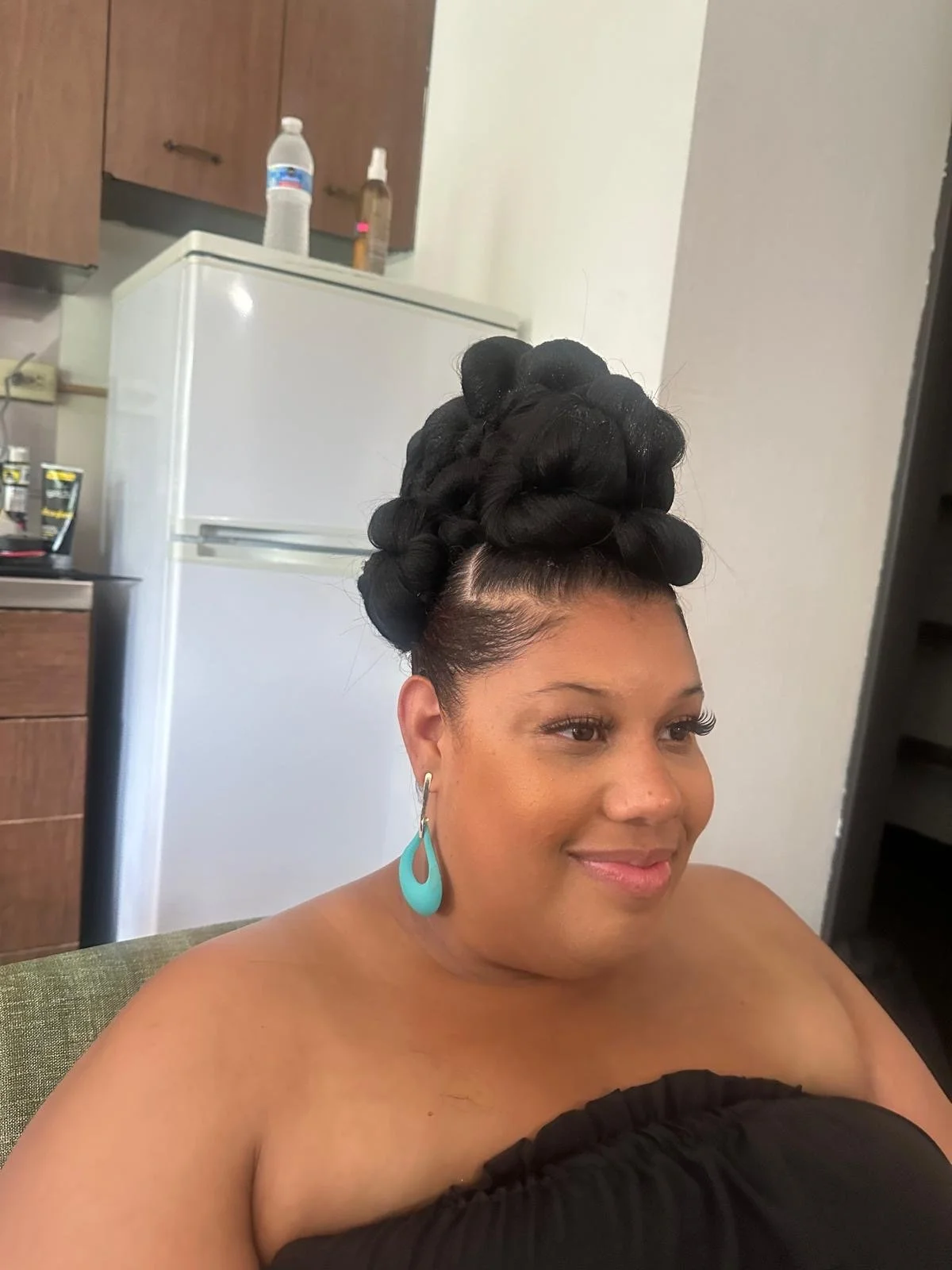 A woman with an elaborate black hairstyling with large rolled buns, wearing turquoise earrings, smiling softly while sitting on a green chair in a kitchen.