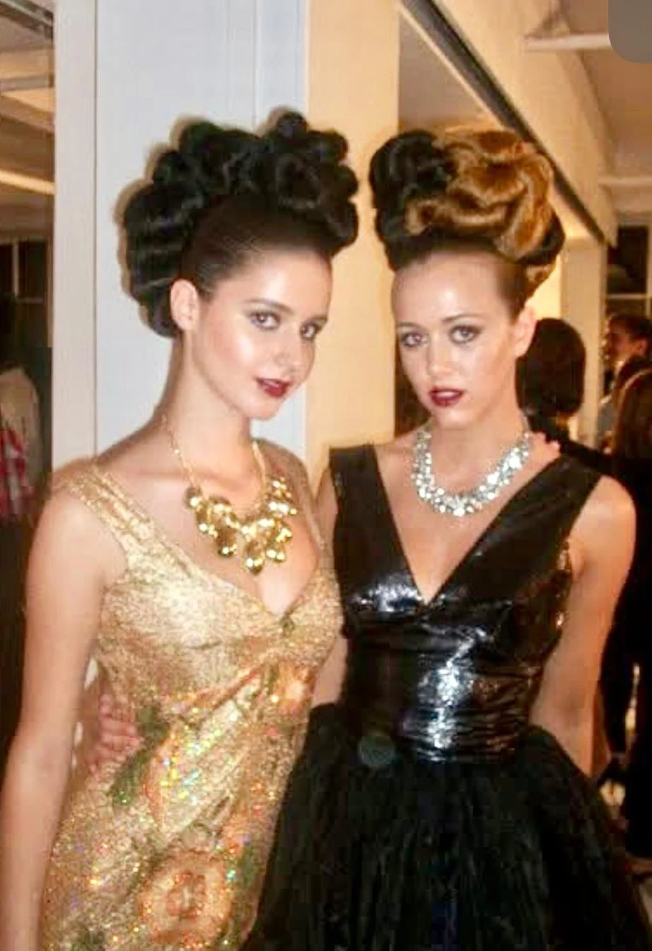 Two women with elaborate updos and glamorous makeup, wearing glamorous dresses and statement jewelry, standing close together at an indoor event.