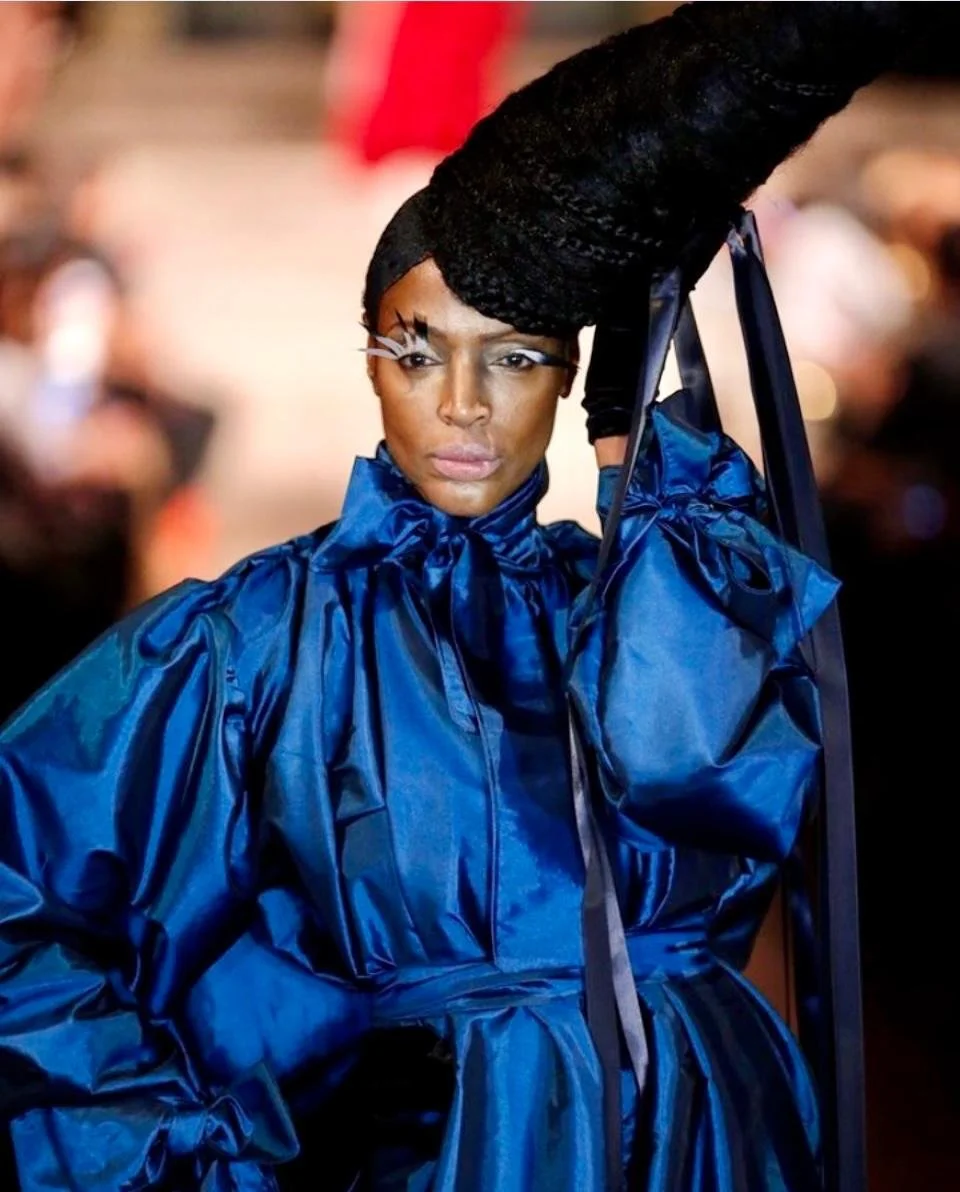 A woman in a striking blue shiny outfit with elaborate black hair styled high, holding a frame or structure around her head, sporting bold makeup with dramatic white eyeliner and accessories.