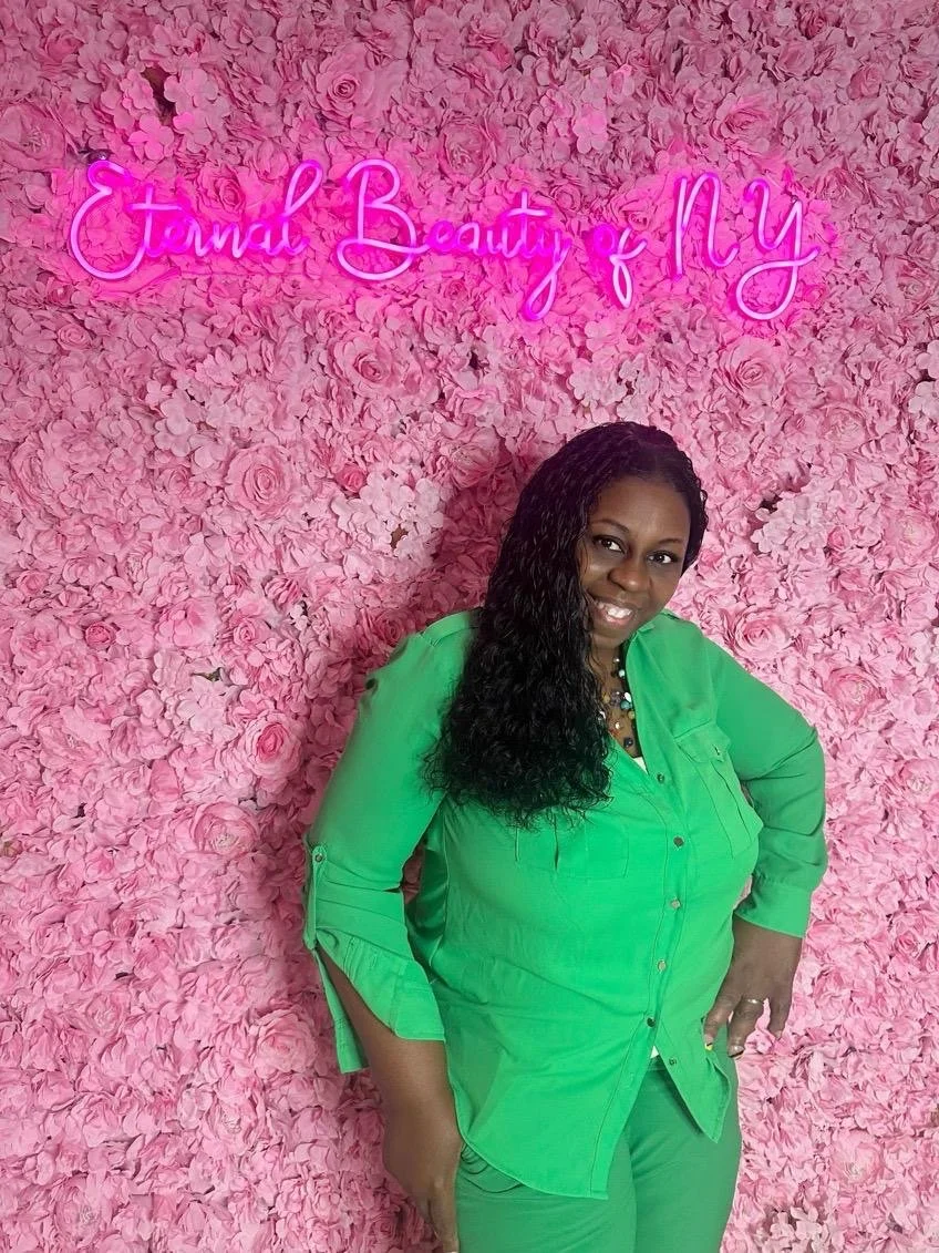 A woman with long curly black hair wearing a bright green outfit, standing in front of a pink floral wall with neon pink sign reading 'Eternal Beauty & My'.