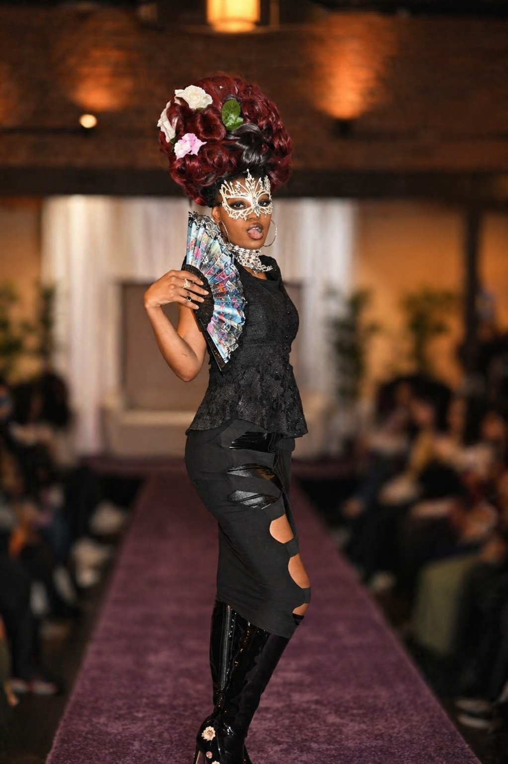 A person walking down a runway wearing a dramatic black lace top, distressed black pants, and shiny black high-heeled boots. They have an elaborate red and black hairstyle decorated with flowers, and they are wearing an intricate white face mask, lar
