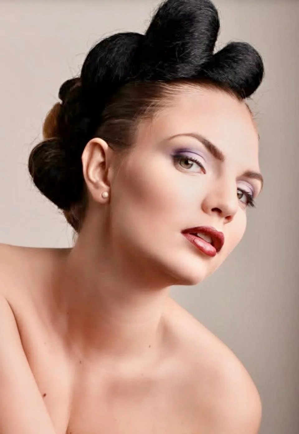 A woman with vintage-inspired rolled and pinned updo hairstyle, wearing earrings, with makeup including purple eyeshadow and red lipstick, looking at the camera with a neutral background.