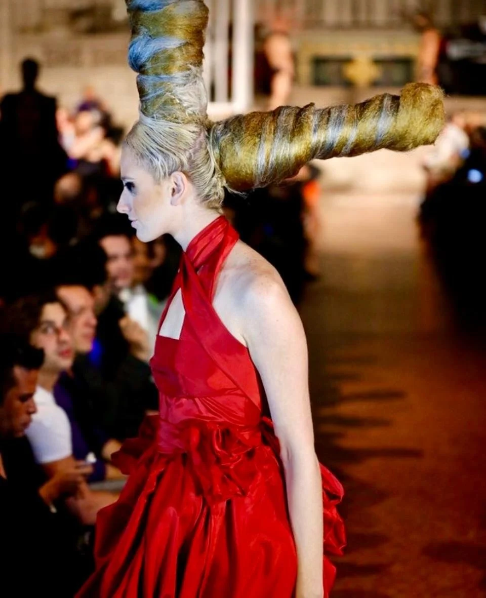 A model with an elaborate gold and silver twisted hairdo on a runway, wearing a red dress, with an audience in the background.