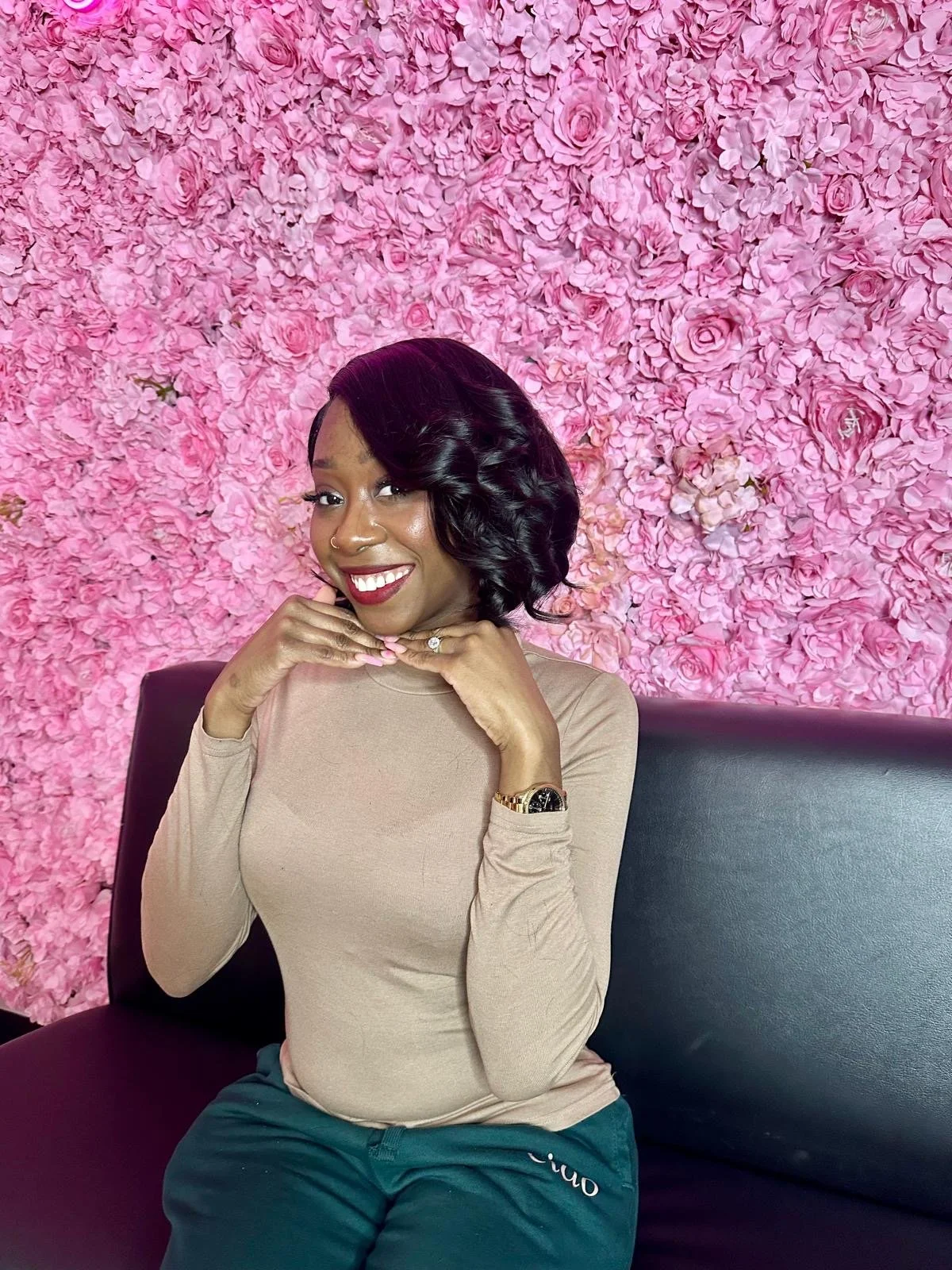 A woman with black, wavy hair sitting on a black bench in front of a pink flower wall, smiling and holding her chin with one hand. She is wearing a beige long-sleeve shirt, dark green pants, a gold watch, a ring, and a nose ring.