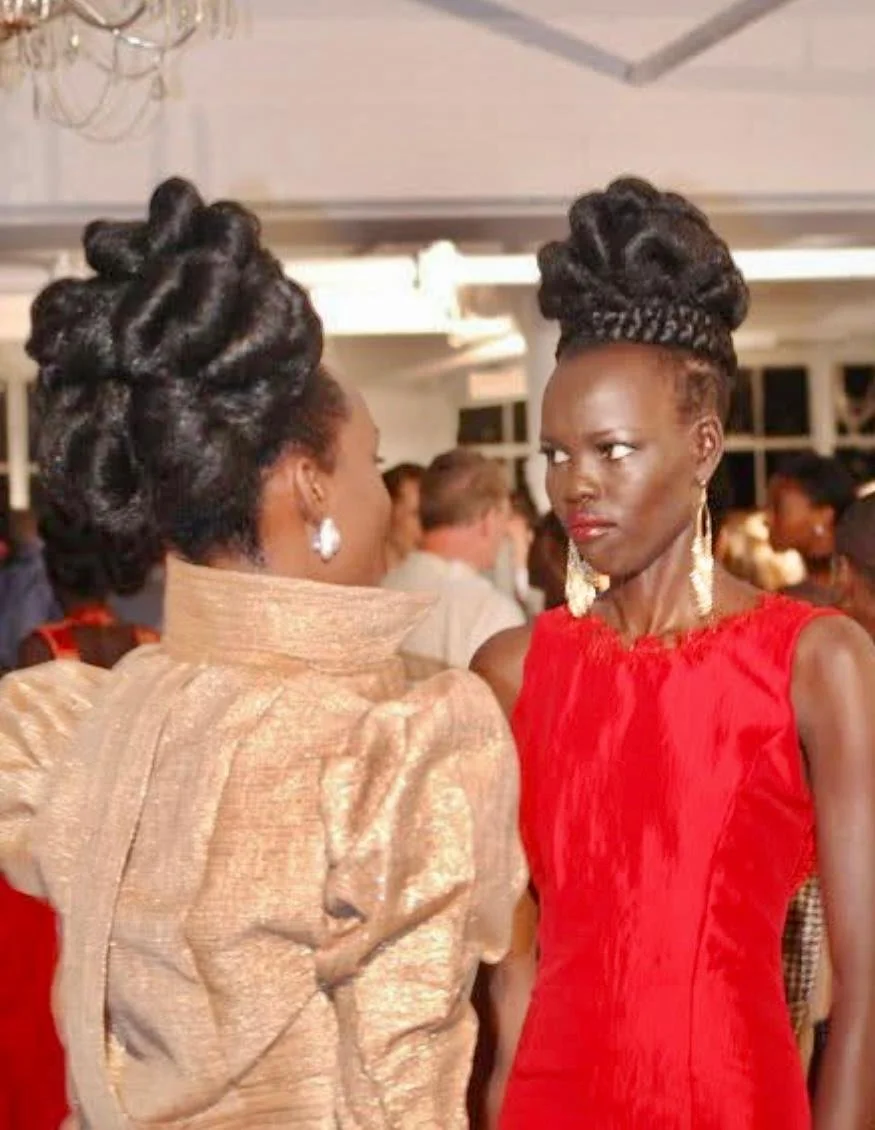 Two women with elaborate updo hairstyles and earrings engage in a serious conversation at a social event, with a crowded background in an indoor setting.