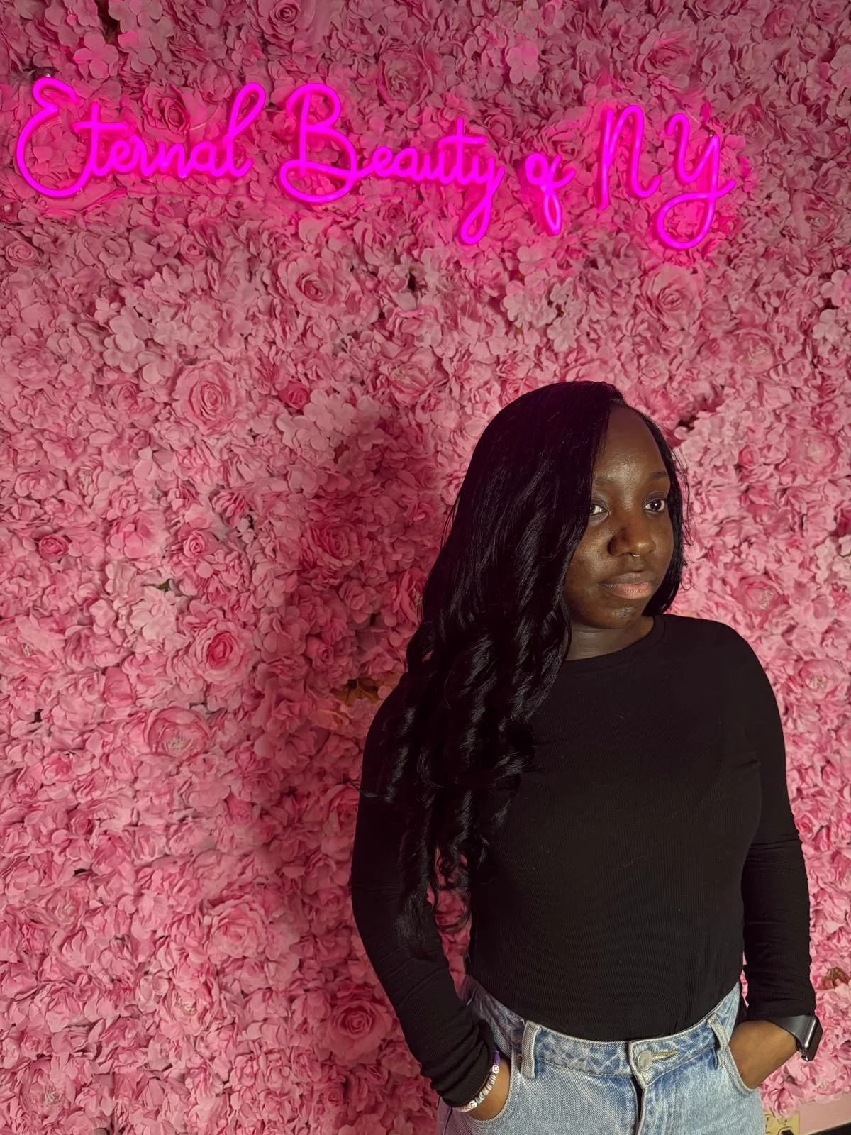 A woman with long black hair wearing a black long sleeve shirt and light blue jeans, standing against a backdrop of pink roses and a glowing pink neon sign that reads 'Terminal Beauty of NY.'