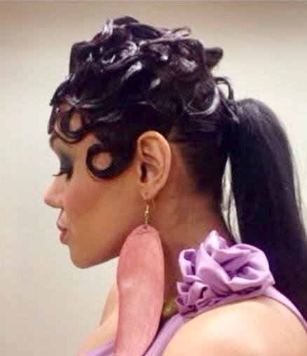 A woman with elaborate curled hair, large pink earring, and wearing a purple top with ruffled details.