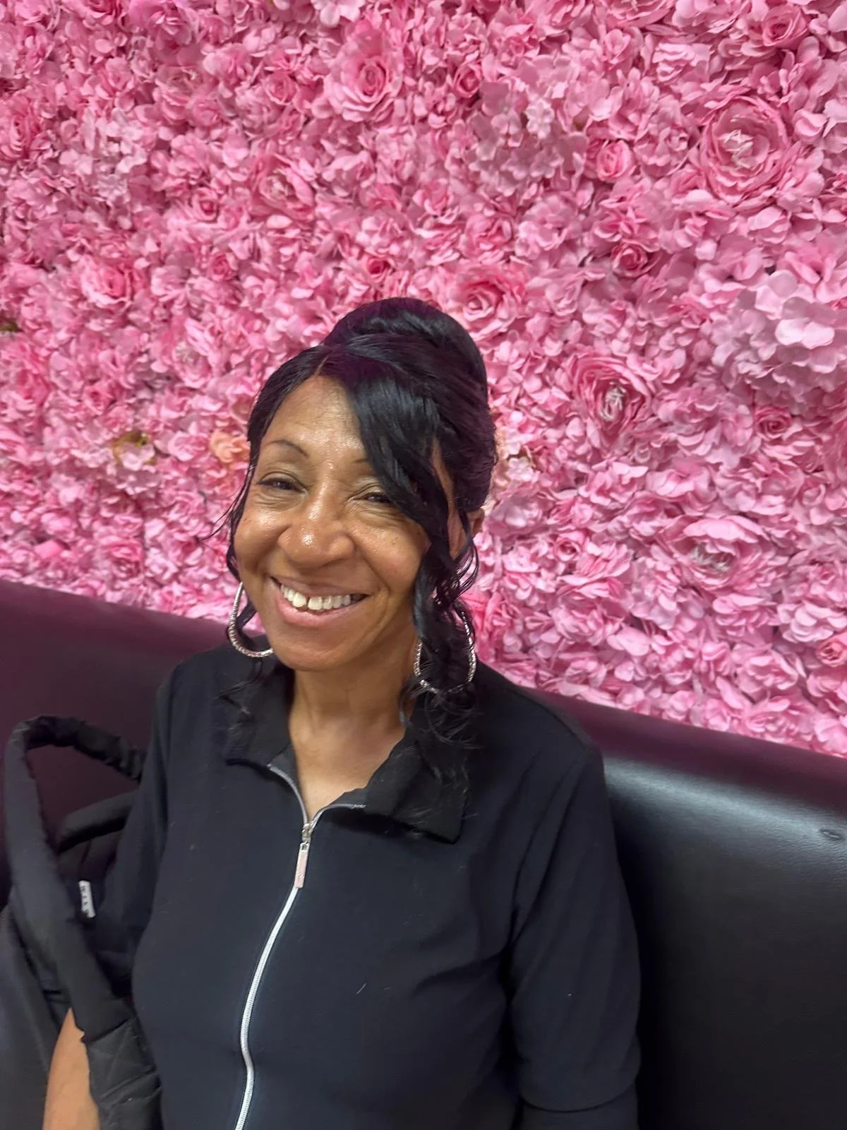 A smiling woman with black curly hair wearing a black zip-up jacket, sitting in front of a wall filled with pink roses.