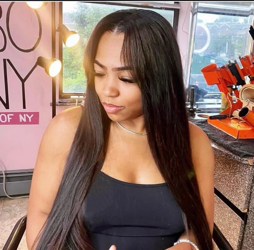A woman with long dark hair wearing a black tank top and necklace, sitting in a cafe with pink walls, large windows, and a work area with orange tools in the background.
