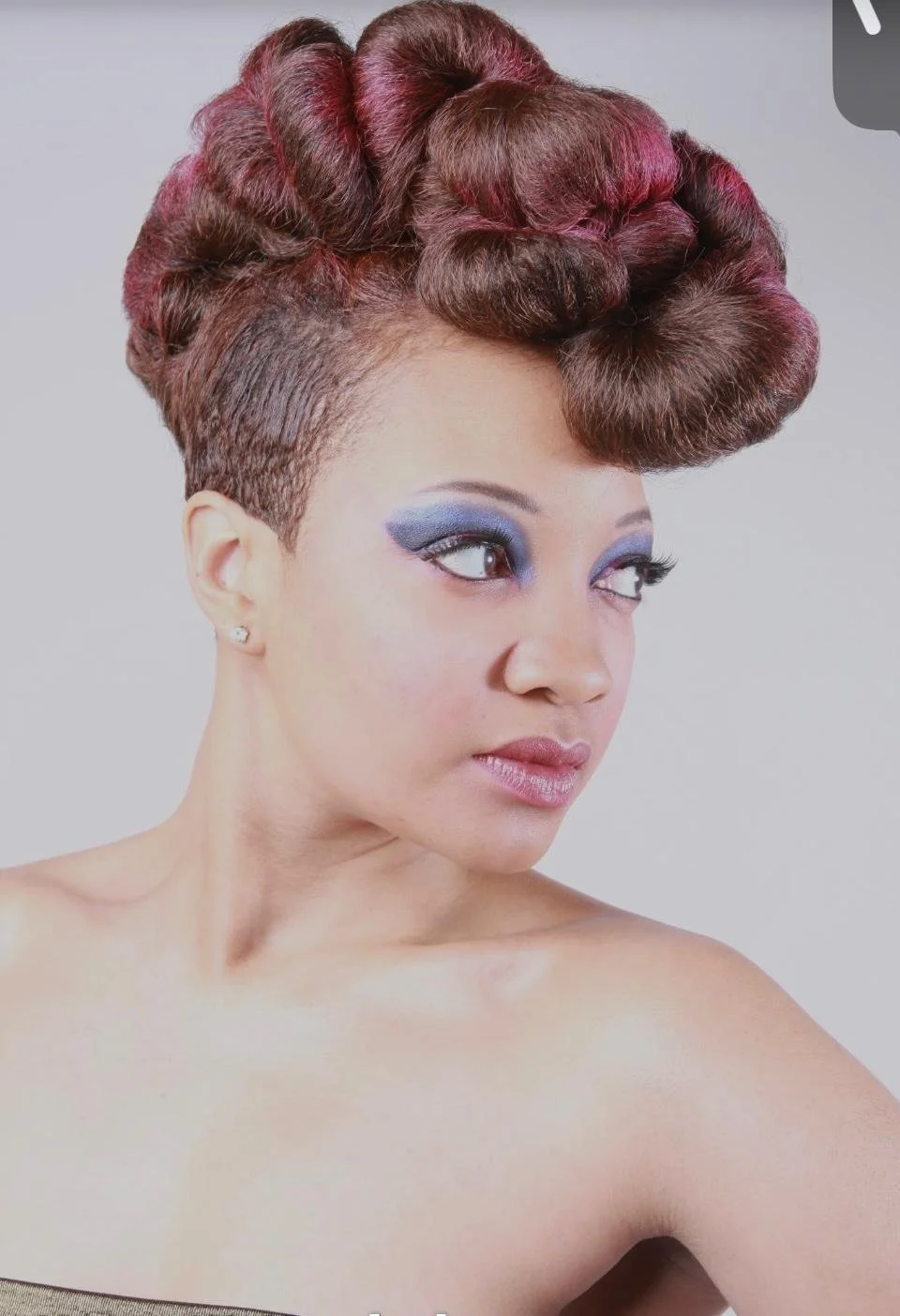 A woman with short, intricate hairstyle with large rolled curls on top and shaved sides, wearing bold blue eye makeup and a small diamond stud earring, looking to the side.