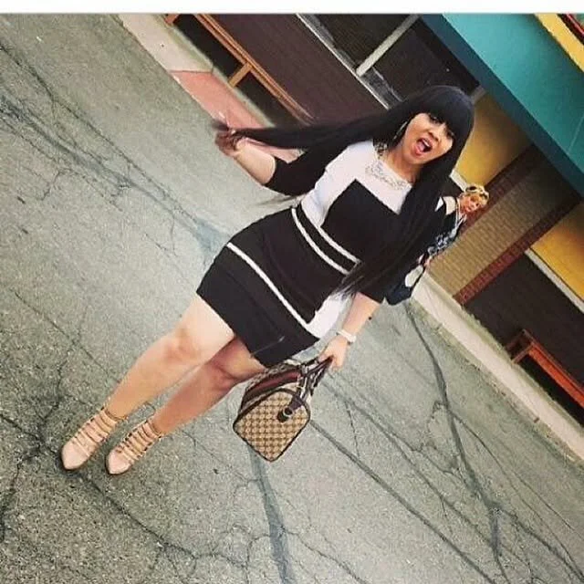 A woman with long black hair and bangs standing on a cracked sidewalk outside a colorful building. She is wearing a black and white striped dress, beige high-heeled shoes, and holding a small patterned handbag. She is make a playful, surprised expres