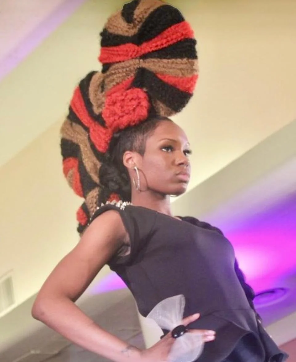 A woman with short hair and hoop earrings stands with her hand on her hip in front of a colorful background. She is wearing a black outfit and a large, elaborate headwrap made of thick, multicolored yarn in red, black, and brown stripes.
