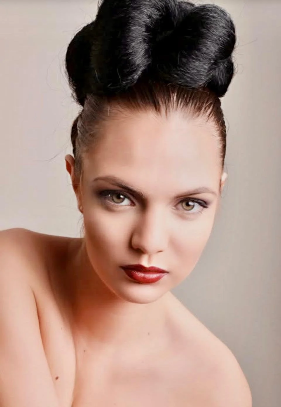 Close-up portrait of a woman with styled dark hair, fair skin, and red lipstick, looking confidently at the camera against a neutral background.