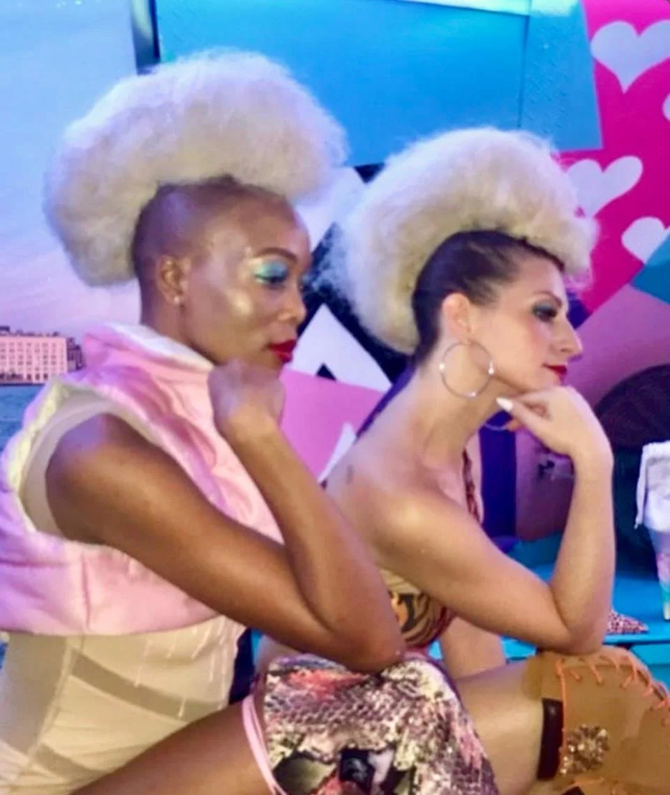 Two women with platinum blonde voluminous hair, sitting side by side at a colorful, heart-themed background. They are dressed in stylish, vibrant outfits, with one wearing a pink top and the other a patterned dress.