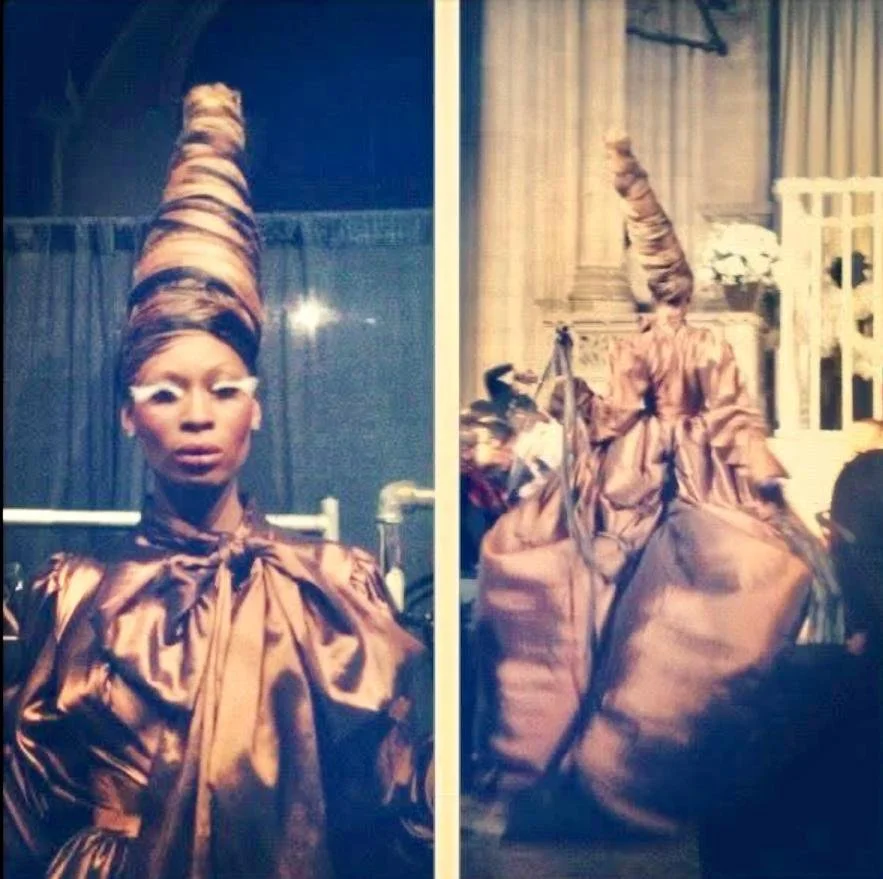 A woman dressed in a shiny copper-colored outfit with a tall, spiral-shaped headpiece, posing indoors in front of a curtain.