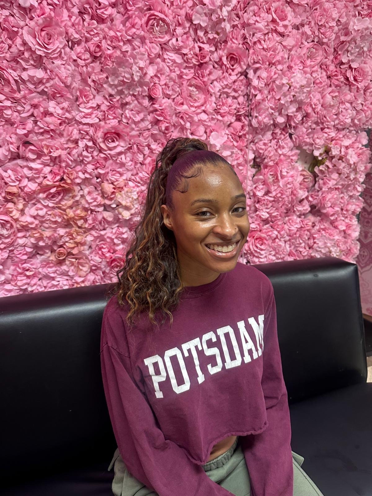 A young woman with curly hair and a high ponytail, smiling, wearing a maroon sweatshirt with white text, sitting on a black bench in front of a pink floral wall.