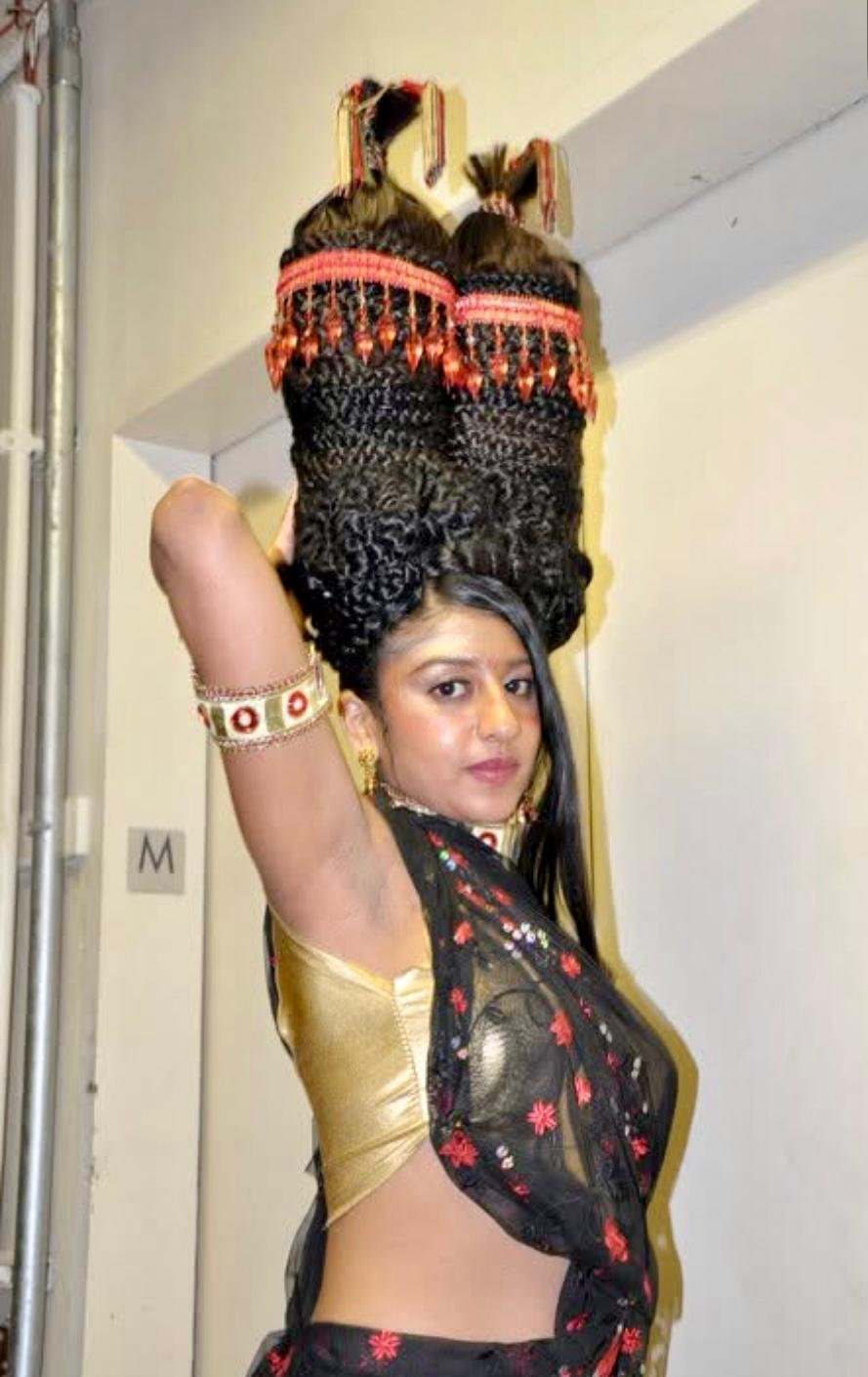 A woman with dark hair wearing a black and gold sari with red floral embroidery, gold jewelry, and a black and red headband, balancing two large bundles of yarn or fabric on her head.