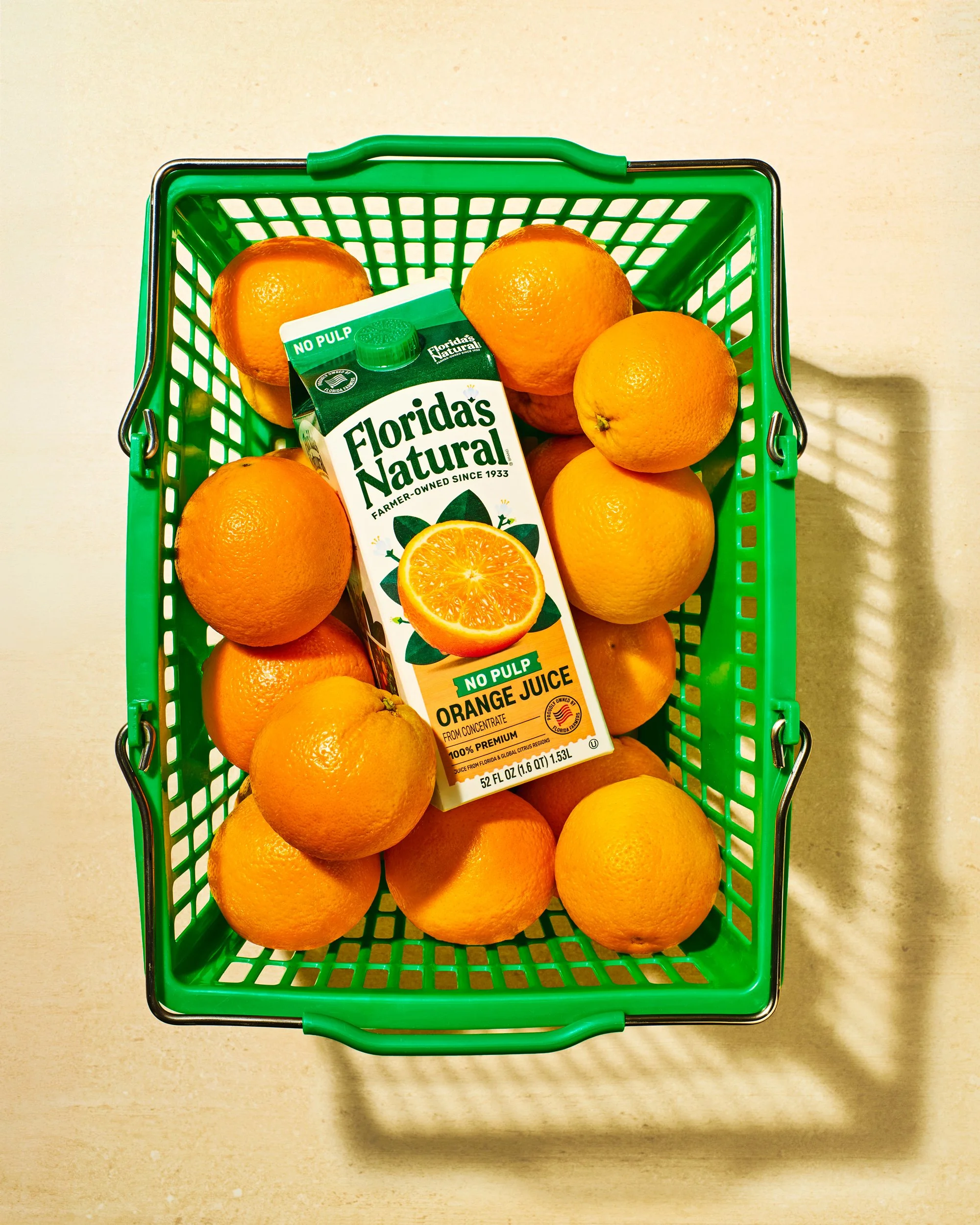 A green shopping basket filled with oranges and a carton of Florida's Natural orange juice on a beige surface.