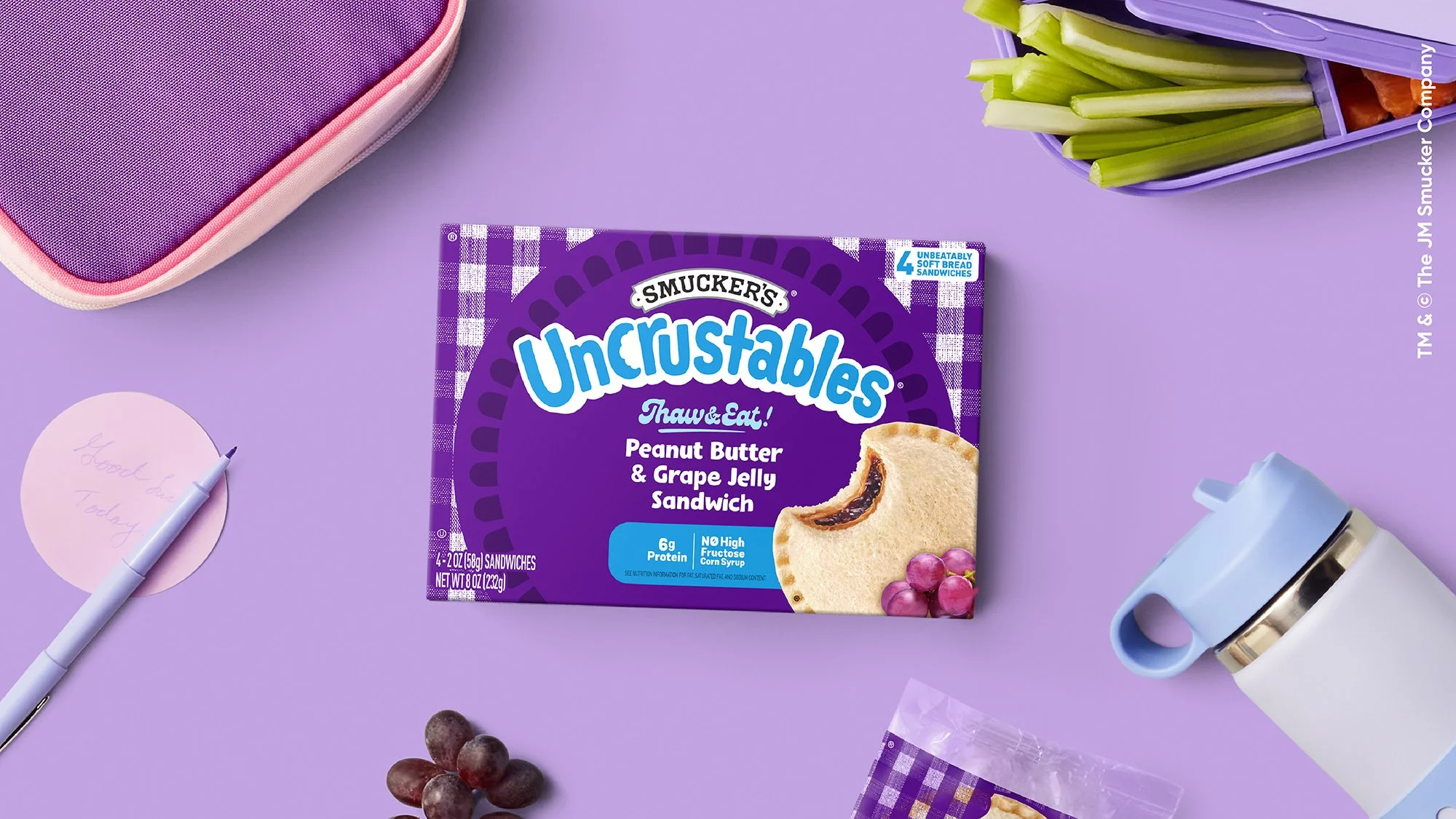 A purple box of Smucker's Uncrustables peanut butter and grape jelly sandwiches on a purple background surrounded by a pink notebook, a white pen, a purple container with celery sticks, grapes, and a blue mug with a tea infuser.