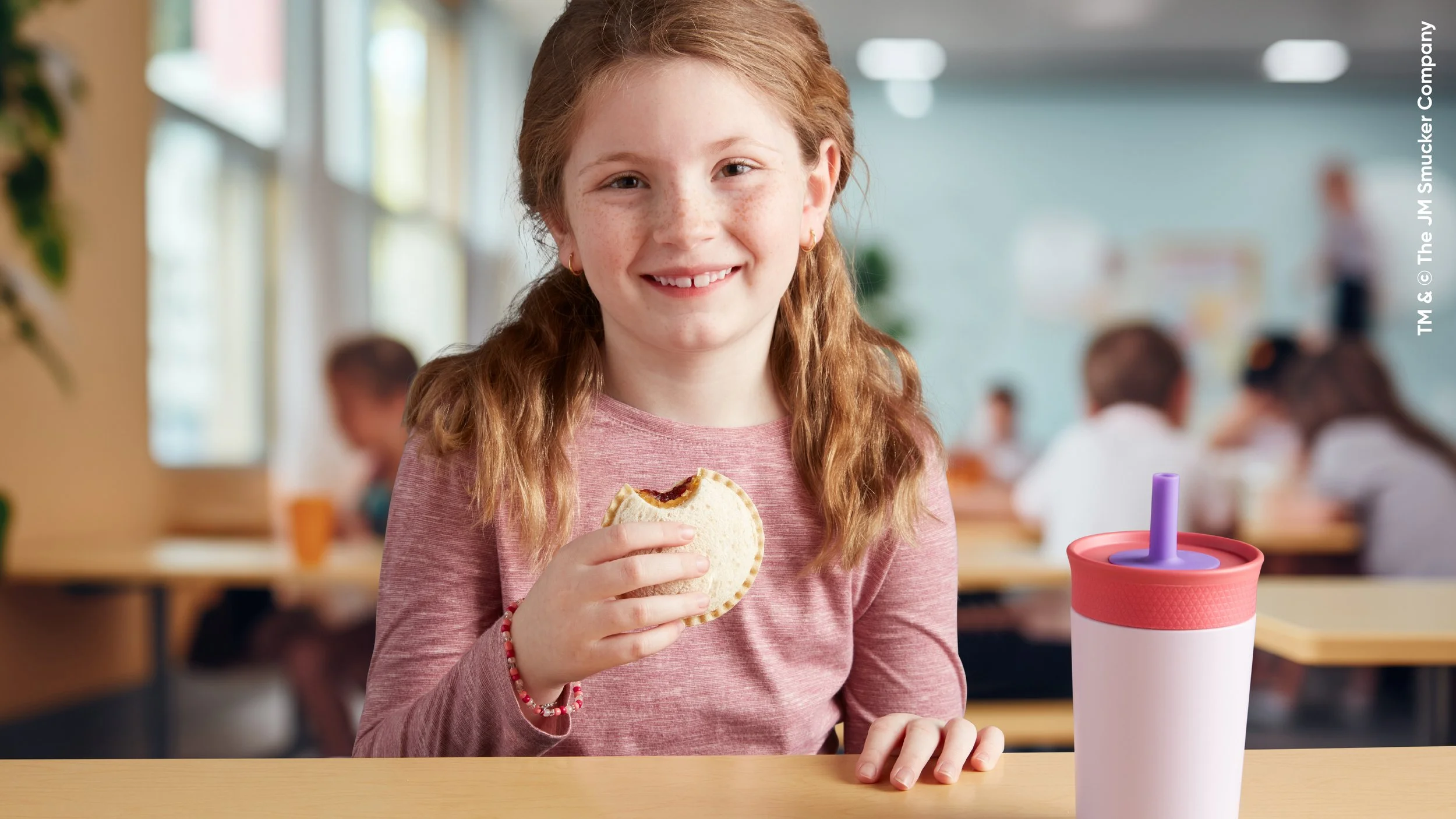 A young girl with red hair and freckles smiling while holding a sandwich in a cafeteria setting. She has a pink shirt and is sitting at a table with a pink and white cup with a purple straw in front of her. In the background, there are other children and large windows.