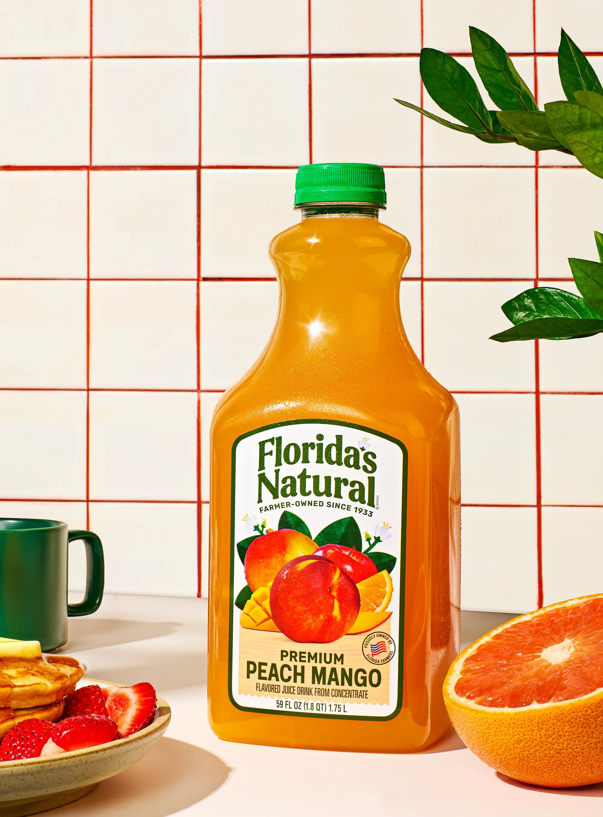 A bottle of Florida's Natural brand peach mango flavored juice with a green cap, surrounded by slices of grapefruit, strawberries, and a bowl of strawberries, set against a white tiled background with red grout lines.