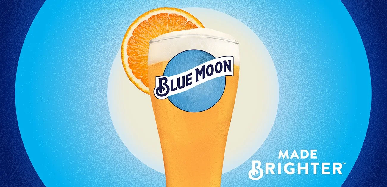 A glass of Blue Moon beer garnished with an orange slice, set against a blue background with the Blue Moon logo and the phrase 'Made Brighter'.