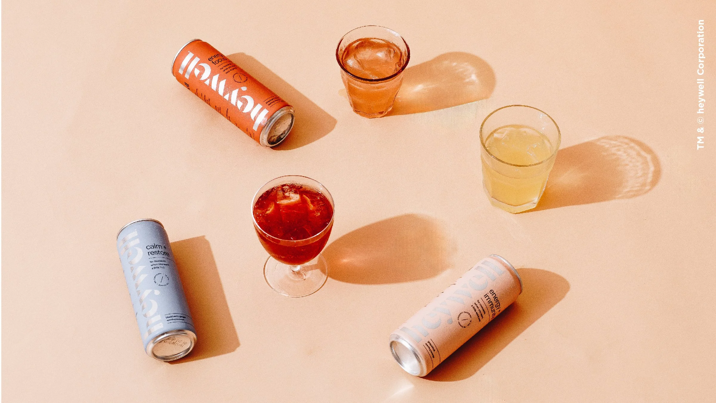 Assorted cans and three glasses with colorful drinks on a peach-colored surface, casting shadows.