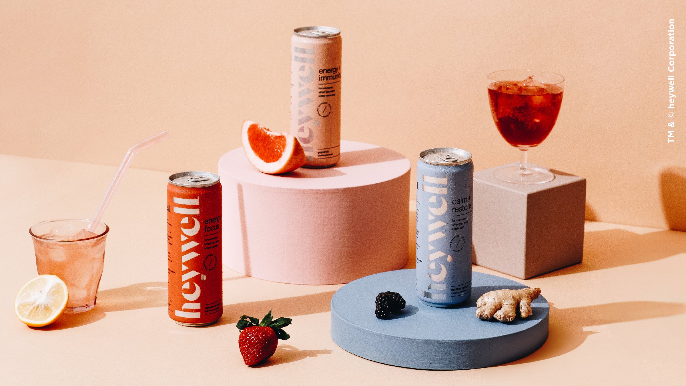 Arrangement of energy drink cans, a glass of pink beverage with ice and lemon wedge, pink and blue circular platforms, and fresh strawberries, blackberries, and ginger on light pink background.
