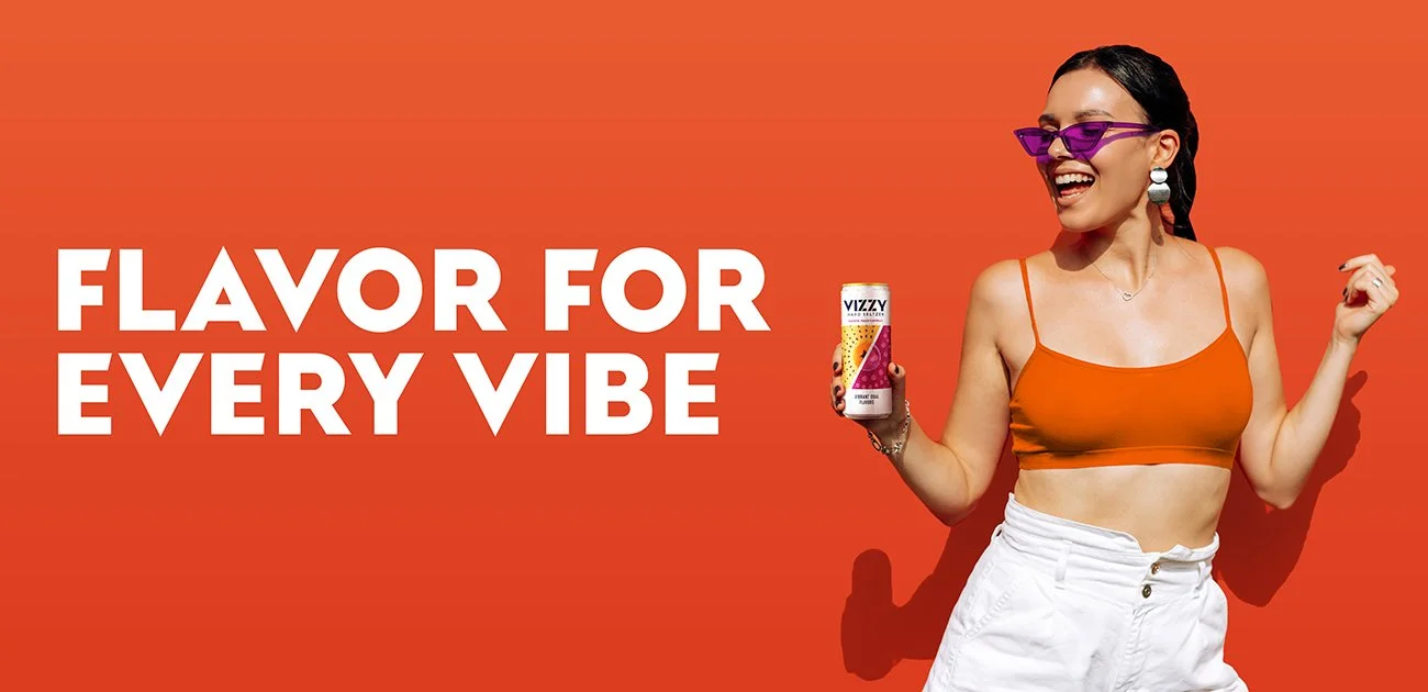 A woman with dark hair, glasses, and earrings smiling while holding a can of Vizzy flavored sparkling water, standing against an orange background with the text "Flavor for Every Vibe."