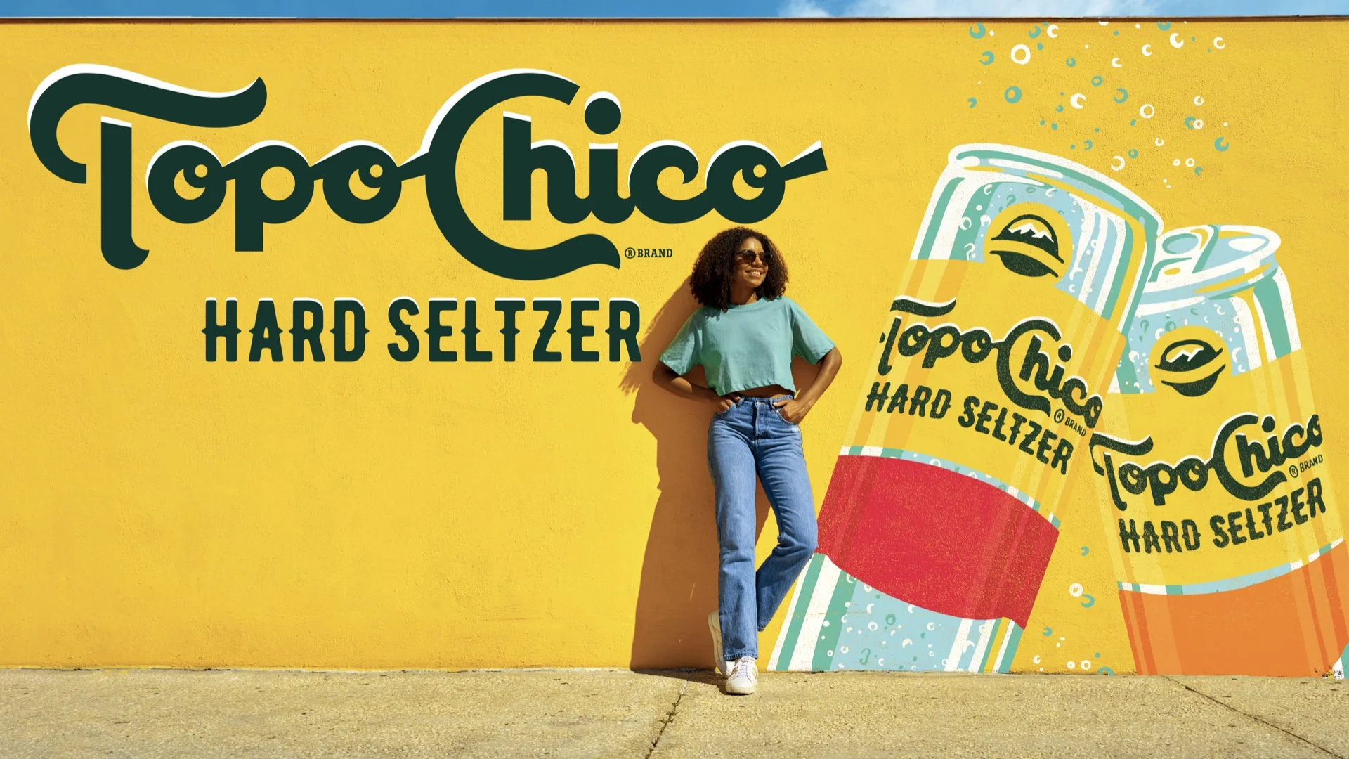 A woman wearing sunglasses, a teal t-shirt, and jeans standing against a bright yellow wall with large graphics of Topo Chico Hard Seltzer bottles and the brand's logo, smiling with one hand on her hip.