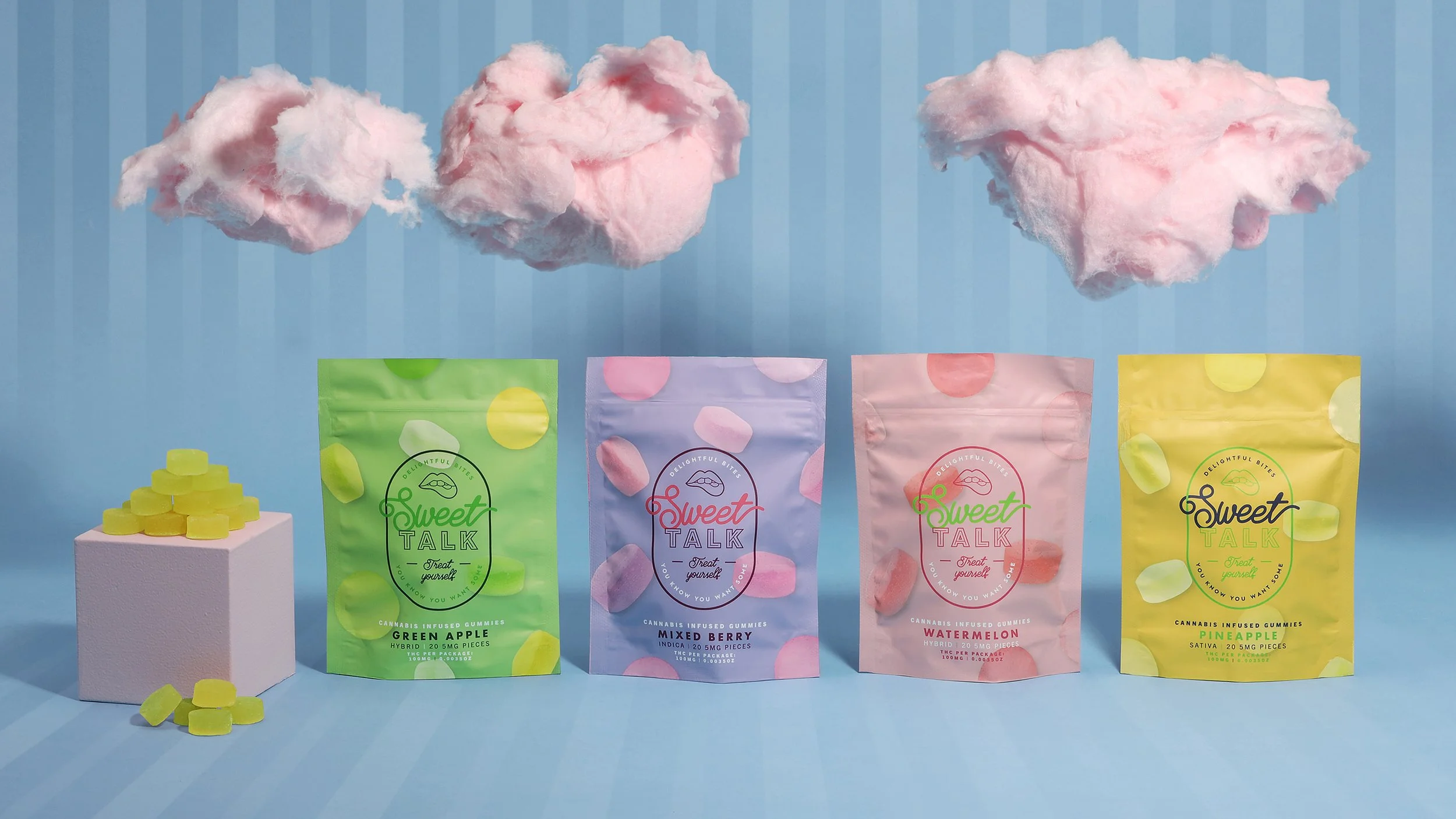 Four colorful cannabis-infused gummy packages labeled 'Sweet Talk' lined up on a blue background, each featuring different fruit flavors: green apple, mixed berries, watermelon, and pineapple. A white pedestal on the left displays a small pyramid of yellow gummies.