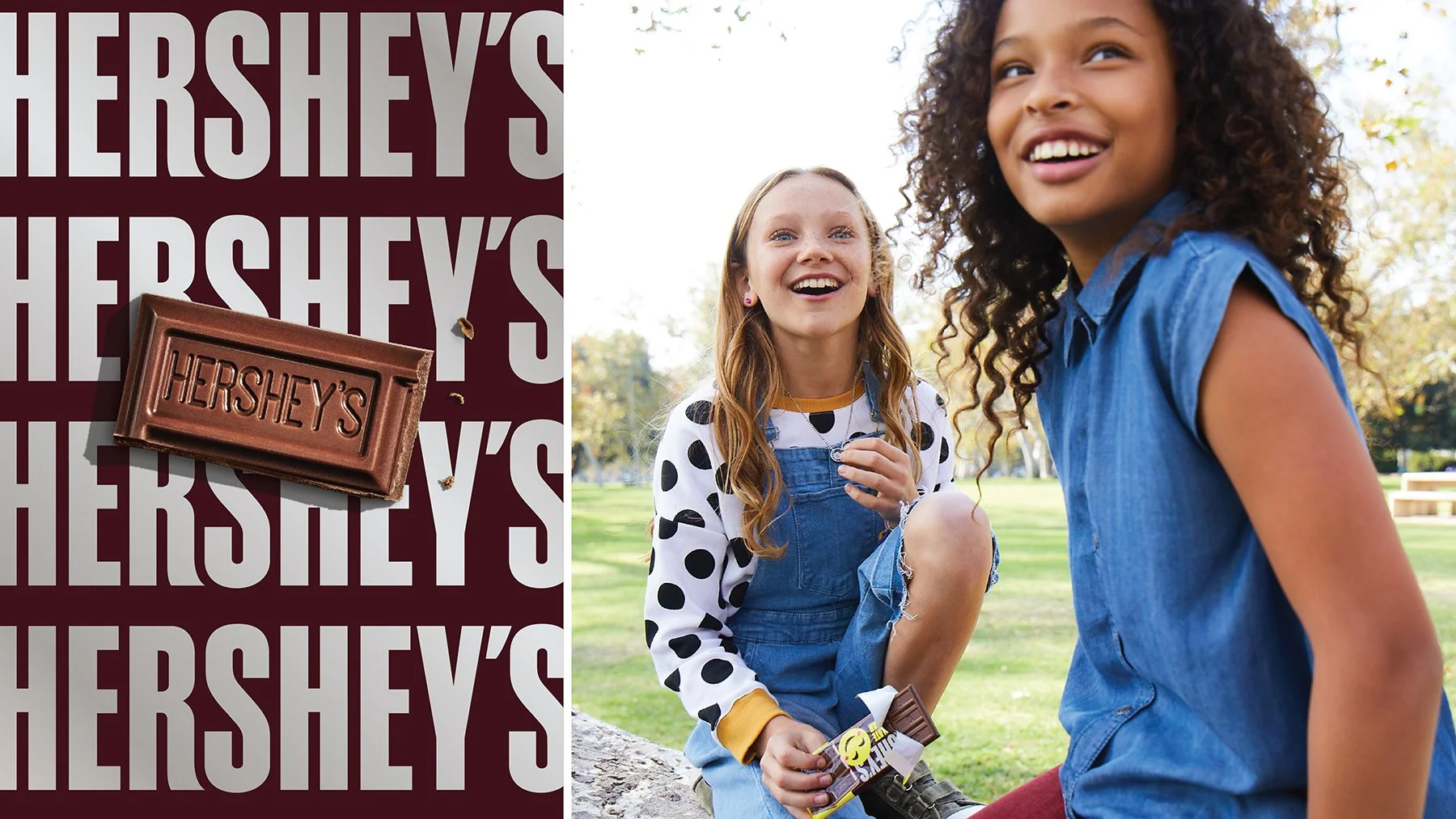 Two young girls outdoors at a park, one with curly hair and a blue denim shirt, smiling, and the other with red hair in a polka dot shirt, holding a Hershey's chocolate bar