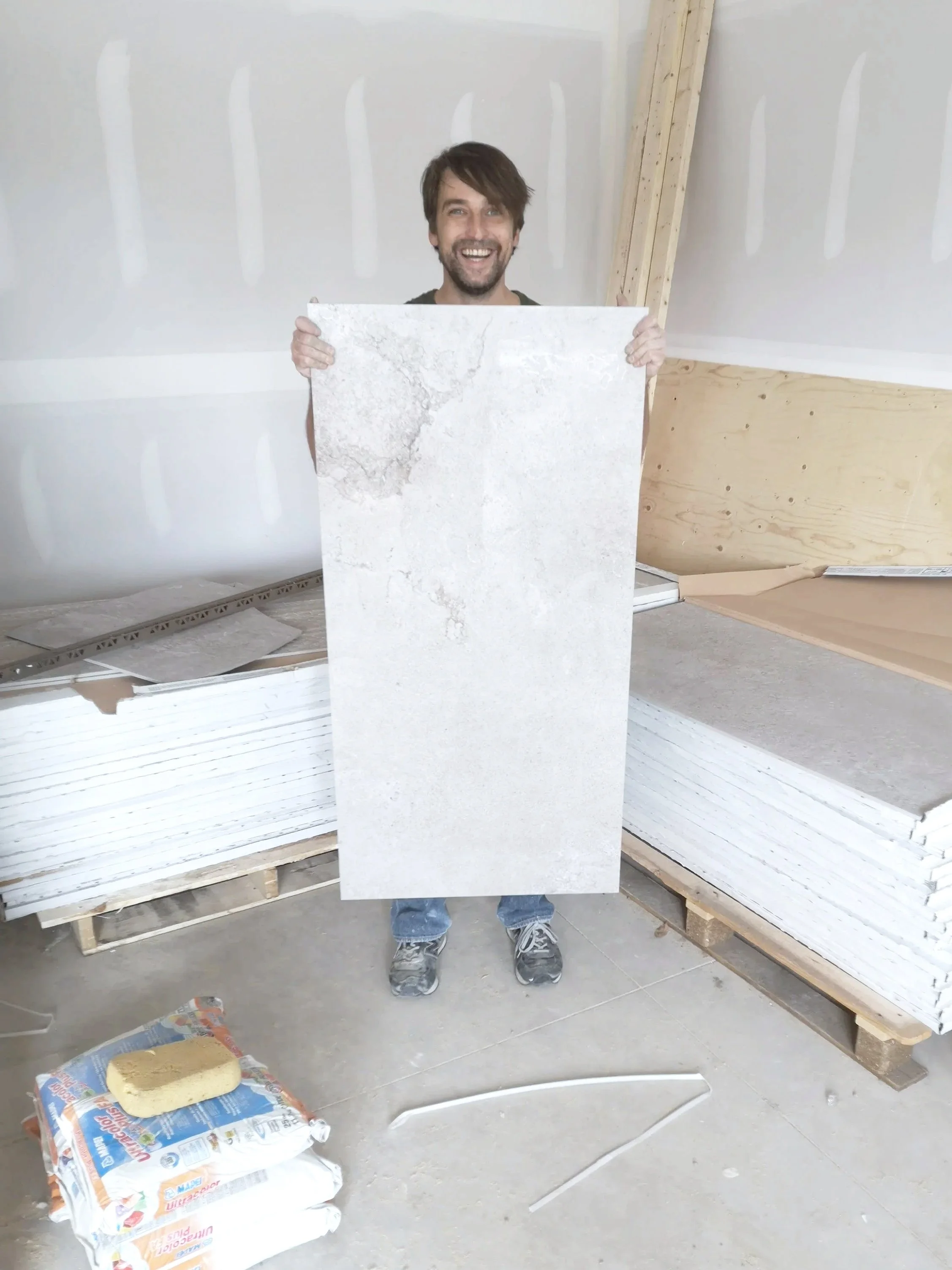 A smiling man holding a large tile in a construction site with drywall and stacks of other tiles in the background.